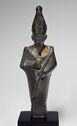 A dark bronze standing figure of a man. Both of his hands are in front of his chest holding long, thin objects. He wears a detailed golden neck piece. He has a long goatee and a tall head piece.