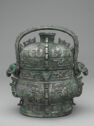 A gray cast bronze vessel with a lid and a middle handle on top of it. It is smaller at the top and wider at the bottom with a thick foot. It is inscribed with detailed, swirling designs all over. The ends of the handle have ram heads coming out.