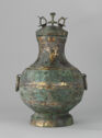 A cast bronze vessel that has a short round body, a tall narrow neck, and a lid that has three pieces standing on it. It has thick bands of green and gold along the sides. The body has two rings attached on either side.