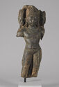 A sculpture of a man standing with his hip leaned to the right. He is wearing a decorated belt, necklaces, earrings, and a crown. On each side of his head are two smaller heads also wearing crowns. His legs below the knees and arms below the shoulders have been broken off.
