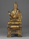 A gilt bronze sculpture of a man seated with one hand raised.