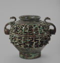 A dark brown/green bronze vessel that has a detailed, overlapping 3D pattern along the sides. The pattern has small inlaid turquoise and a white material. There are two curved serpents for handles on both sides. The top lip is short and flares out.