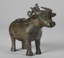 A cast bronze buffalo-shaped vessel that stands on four legs. It has an olive-green patina and its head has a sloped, flat snout, wide eyes, teardrop shaped ears, and horns that have an engraved pattern. There is a curved handle on its back behind its head and an indented line around its back that shows where the lid separates.