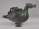 A dark green cast bronze vessel in the form of a duck. It stands on its two feet with its head up and facing the right. It has a wide lid on its back that is attached by a chain. The entire piece is inscribed with a swirling pattern and fine lines that mimic feathers along the body.