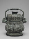A black cast bronze vessel with a lid and a middle handle on top of it. It is smaller at the top and wider at the bottom with a thick foot. It is inscribed with detailed, swirling designs all over. The ends of the handle have animal-like heads coming out.