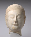 A white marble sculpture of a head of a person starting at the neck. The face is round with small lips, nose, slim eyes, and thin, curved eyebrows. Their hair is short with swirled details.