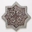 A flat, lusterware tile that is in the shape of an eight pointed star. It is colored a dark brown/grey with a white, patterned band framing the inside of the star shape. White lines make a symmetrical, swirling floral pattern in the center of the piece.