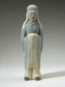 A gray earthenware sculpture of a man standing upright. He is wearing a sleeved garment that covers his entire body with his feet poking out and a draped headpiece. His hands are together in front of his chest, hidden by his sleeves.