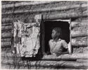 A black-and-white photograph portrays a young Black boy gazing out of the open window of a log cabin.