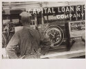 A black-and-white photograph portrays a man with hands on hips facing a bank’s storefront window. 