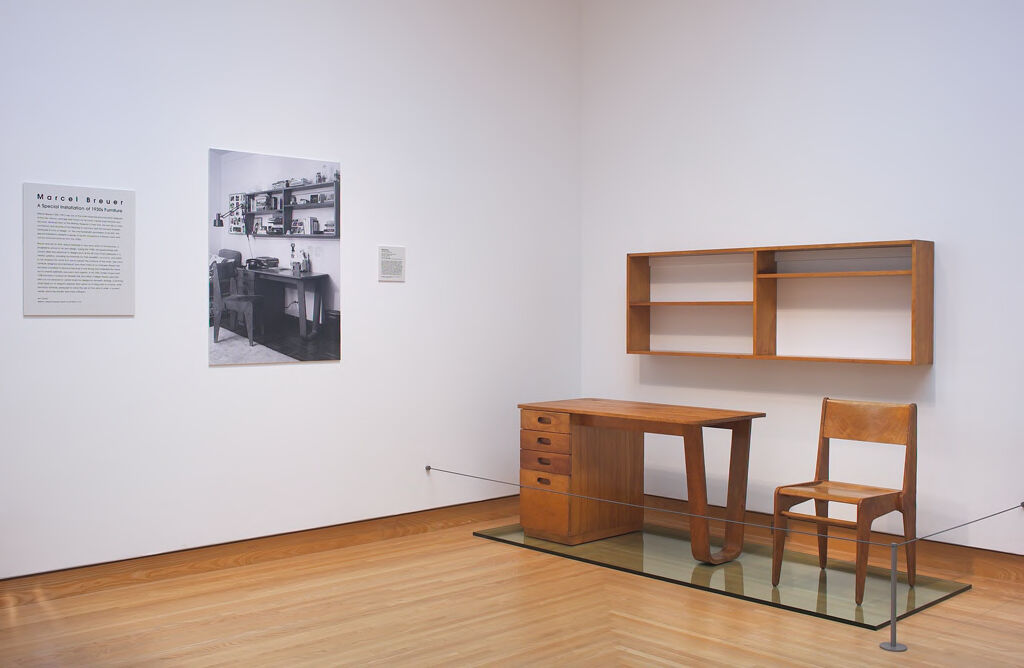 An installation photograph shows a corner of a white-walled room with a display of three pieces of wood furniture against one wall.