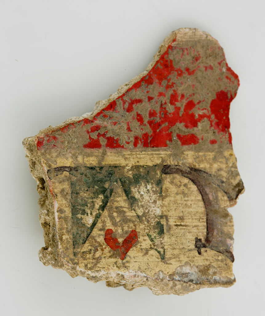 This plaster fragment is unevenly shaped and is partly covered in dirt. The top half is bright red. The lower half is light brown and is decorated with darker-colored geometric shapes and a small red ornament in the form of a heart.