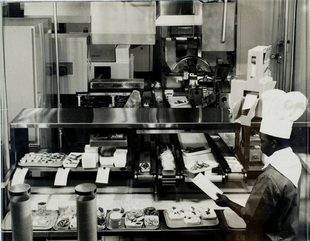 World's First Fully Automated Restaurant (Sixth Of Eight): The Assembly Area Of The Amfare System Finds The Attendant--The Only Person Involved In The Operation Of The System Other Than The Operator Of The Orbis Control Unit--Matching Food Items With An Order Check Dispensed By The Printer On Top Of The Counter Behind His Chef's Hat.  Once Loaded Trays Are Placed On A Converyor Belt.