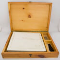 An open, shallow wooden box with a metal clasp, containing a white photo album in one compartment and two smaller objects in their own divided compartments.