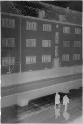 [Reflection Of Apartment Building On Wet Street And Two People Walking]