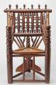This photograph shows an ornate wooden turned armchair with a triangular seat supported by three legs and reinforced by a wooden fin attached to the back leg. The two front arms of the chair have turned details above seat height and are rounded at top. At the pointed back of the triangular seat are a total of nine carved rods that support the upper back. Four thick spindles connect the upper arms to the backrest. Many rods and roundels make up the back of the chair, which is topped with six vertical elements.
