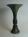 A dark green-grey cast bronze vessel that flares at the bottom and the top. The middle is long and narrow. The whole object is decorated with finely engraved lines that make a pattern except for triangle-shaped areas on the top.