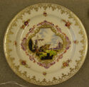 A white plate with an ornate gold design around the edge. Purple flowers circle the plate's indentation. The center features a ship against a sky, in a cartouche that is bordered in purple and gold.