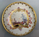 A white plate with an elaborately decorated rim in gold. The center of the plate has a painted harbor scene surrounded in purple and gold decoration.