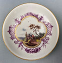 A white plate with a gold rim. A painted of scene of a figure and donkey dominates the center, with a four lobed edge. Purple and gold decoration surround the scene.