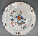 A white plate decorated in blue, yellow, red, and green flora and fauna. The center has a scene featuring baskets, while the edge of the plate has an irregular scalloped edge.
