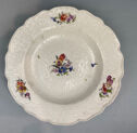 A white plate with scalloped edges and a design in relief. Painted decoration featuring three springs of flowers around the rim, and one in the center.