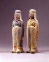 Two gray earthenware sculptures of men standing upright side by side. They are both wearing sleeved garments that cover their entire bodies with their feet poking out and draped headpieces. Their hands are together in front of their chests.
