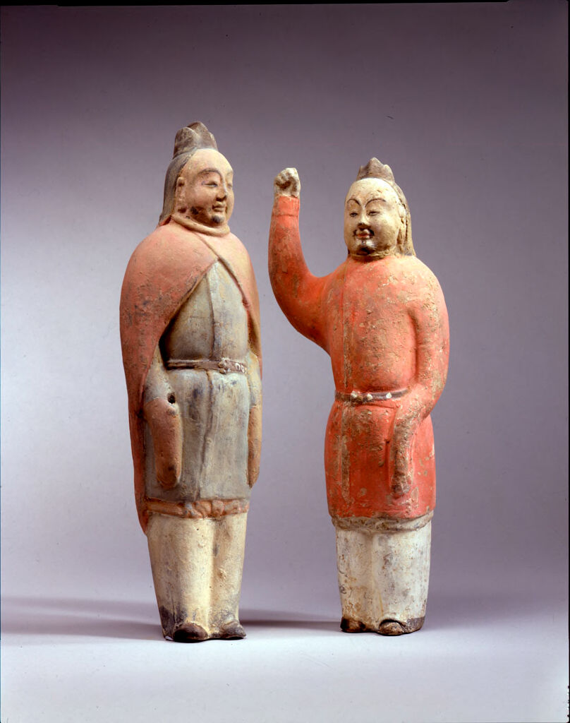 Two Standing Male Figures, Each Wearing A Triple-Peaked Hat With Protective Neck Flap, One Figure Wearing A Cape, His Hands At His Side, And The Other With His Right Arm Raised, His Left At His Side