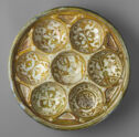 A circular lusterware dish that has seven smaller circular cups within it. It is colored mustard yellow with many painted white details within the smaller cups.