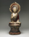 A marble sculpture of a figure sitting cross-legged on a detailed pedestal and wearing a robe. There is a round, detailed form behind their head. There is faded red coloration throughout the sculpture.