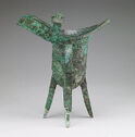 A bright green-grey cast bronze vessel that stands on three narrow, pointed legs, has a tall body, and a top lip that protrudes out. The body is decorated with a finely engraved pattern.