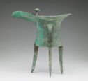 A bright green-grey cast bronze vessel that stands on three thin, pointed legs, has a wide body, and a top lip that protrudes out on both sides. The body is decorated with a vague engraved pattern and has a handle on the side.