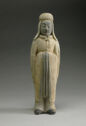 A gray and white earthenware sculpture of a man standing upright. He is wearing a sleeved garment that covers his entire body with his feet poking out and draped headpieces. His hands are together in front of his chest, hidden by his sleeves.