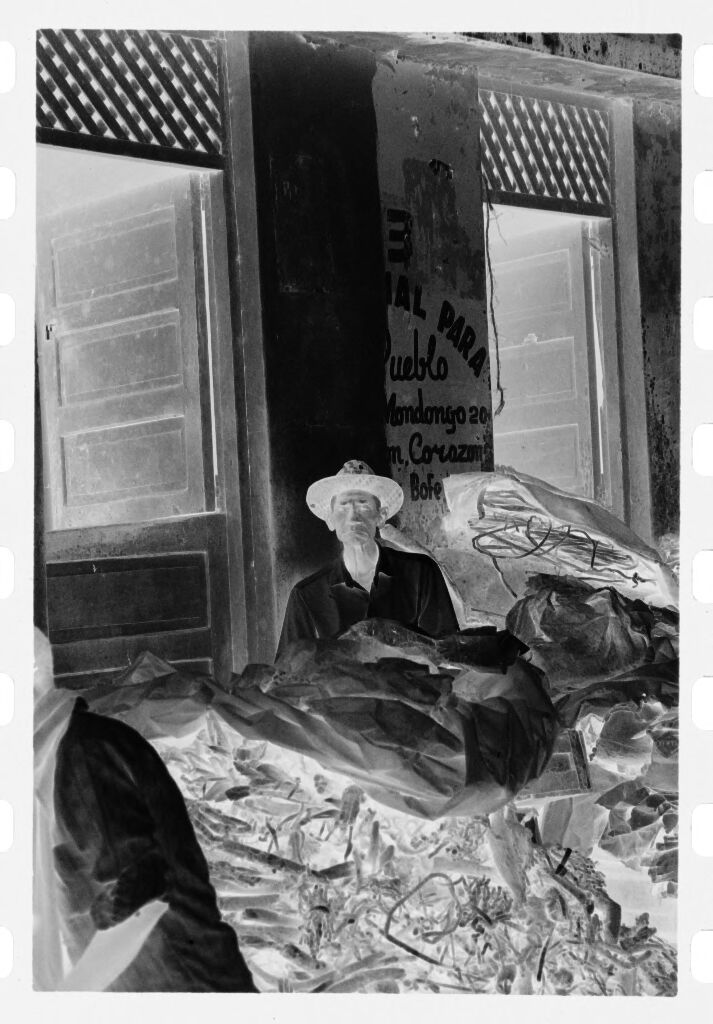 Untitled (Man Sitting In Front Of Market Behind Pile Partially Covered With Tarp)