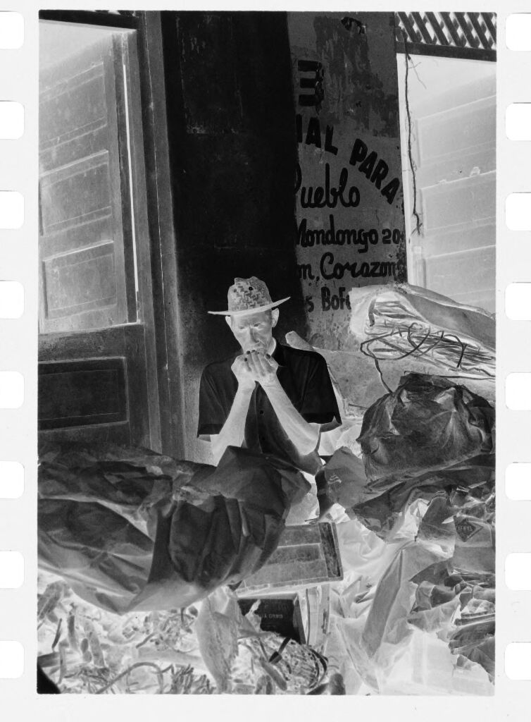 Untitled (Man Sitting In Front Of Market Behind Pile Partially Covered With Tarp)