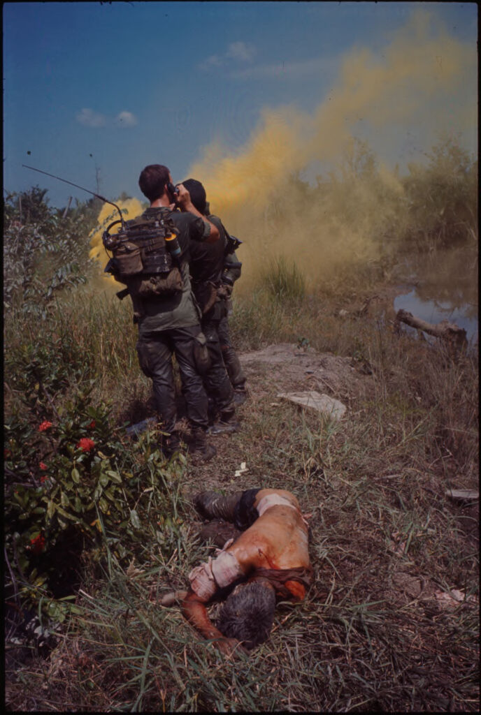 Untitled (Soldiers Standing In Field With Wounded Soldier Lying On The Ground, Yellow Smoke In Background, Vietnam)