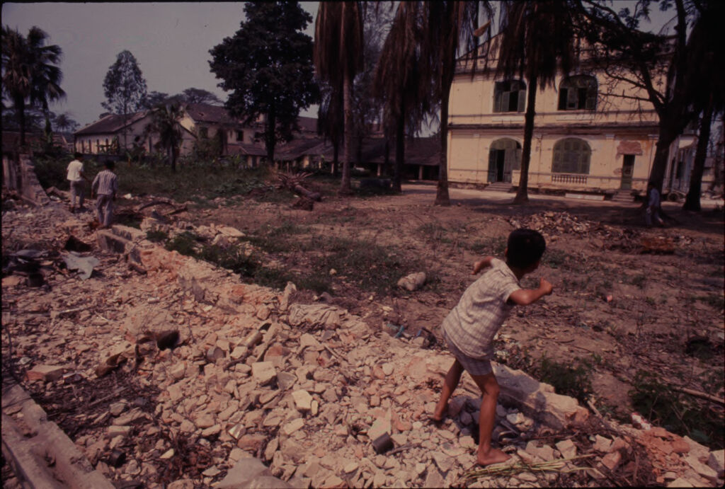 Untitled (Young Boy Throwing Rocks From Rubble Pile Into Field Behind Buildings, Hue, Vietnam)