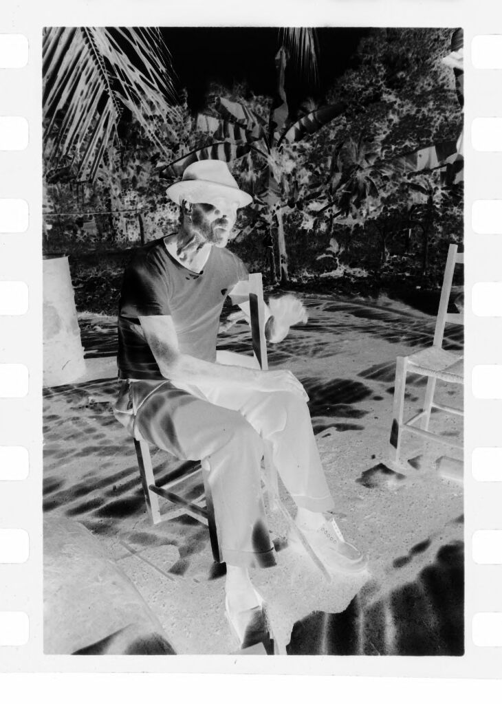 Untitled (Man In White Shirt And Fedora Sitting On Chair Beneath Palm Tree)