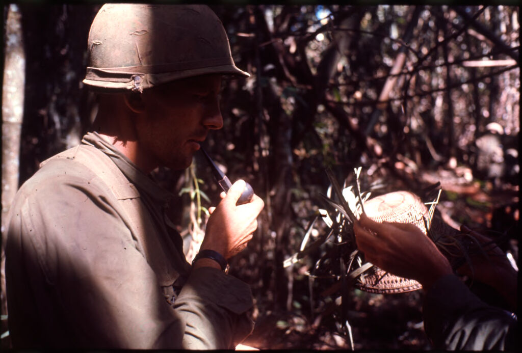 Untitled (Soldier Smoking Pipe Looking At Branch-Camouflaged Straw Hat, Central Highlands Near Dak To, Vietnam)