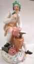 Painted porcelain figure of a young man seated at an anvil with a hammer in his upraised hand.